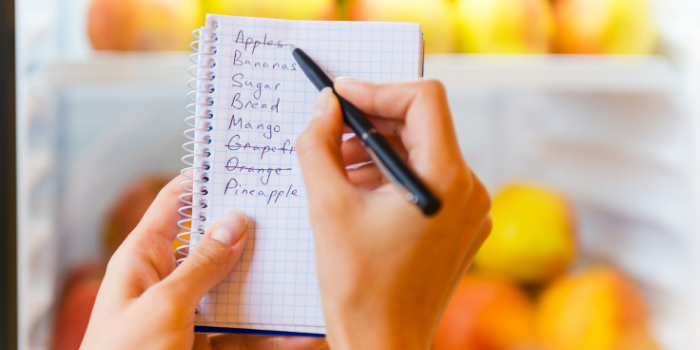 A person holding a pen in their right hand and a shopping list in their left with the words Apple, Bananas, Sugar, Bread, Mango, Grapefruit, Orange and Pineapple on it.