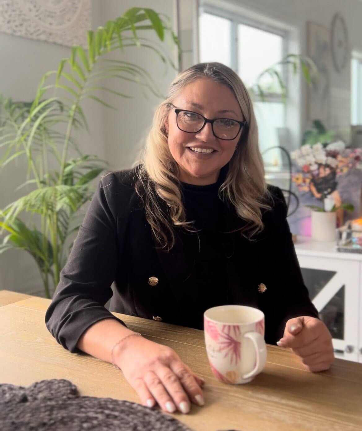 Caroline, a MyBudget client, is sitting at her dining table while holding a white and pink mug in her left hand side while her other hand rests on the table. She is looking directly at the camera and smiling. She is wearing a black top. In the background, there is a medium-sized mirror and plants around it.