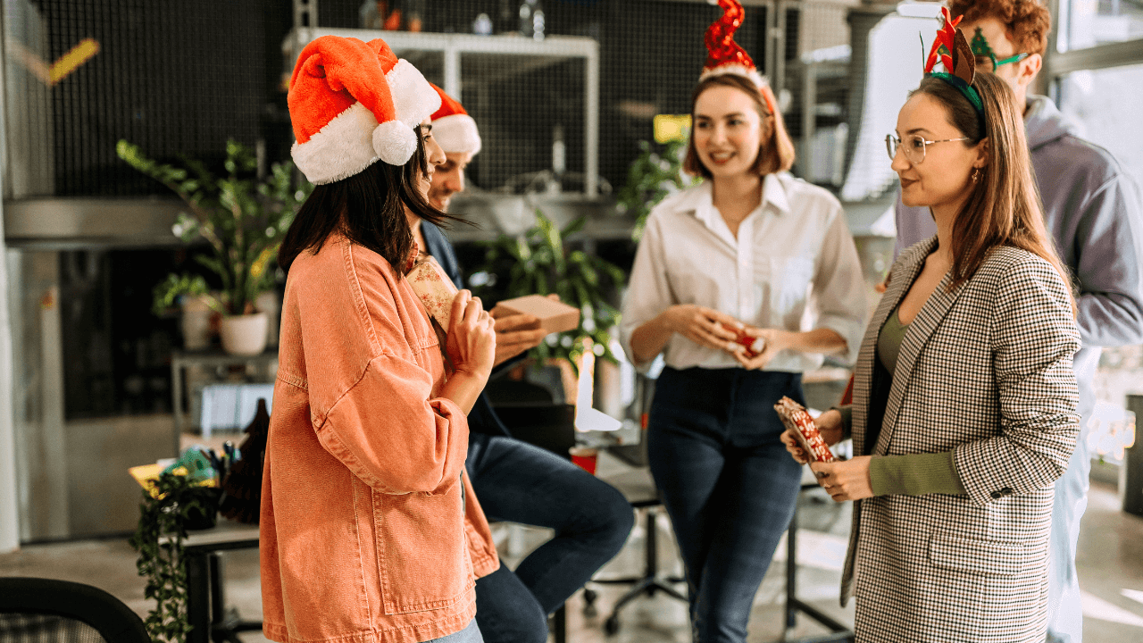 This image showcases a festive office environment with colleagues exchanging gifts while wearing Christmas-themed accessories like Santa hats and reindeer antlers. The scene highlights the joy and celebration of holiday gift-giving.