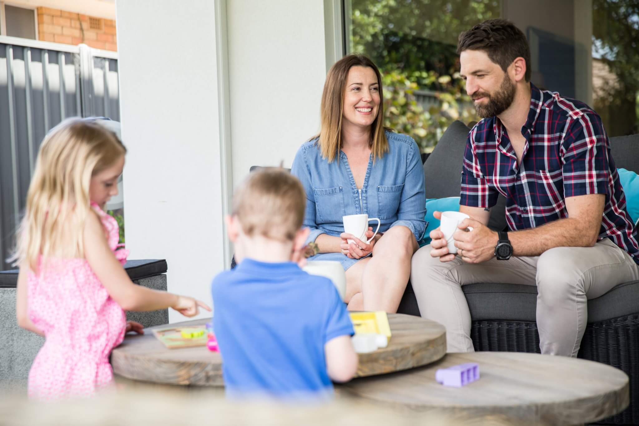MyBudget clients Bec and Jarrad enjoying a coffee outdoors while their kids play, highlighting family time and the importance of budgeting for financial goals.