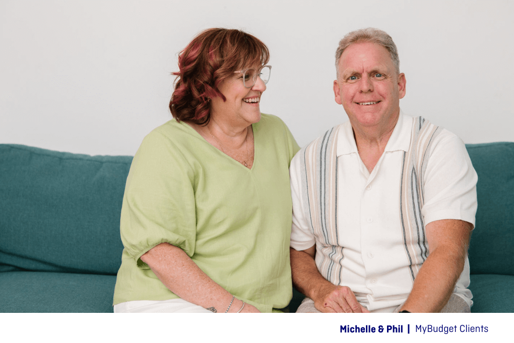 Michelle and Phil, MyBudget clients, high-five and laugh together in front of a mint green backdrop, celebrating their financial success as shared on the MyMoney MyStory podcast with Tammy Barton.