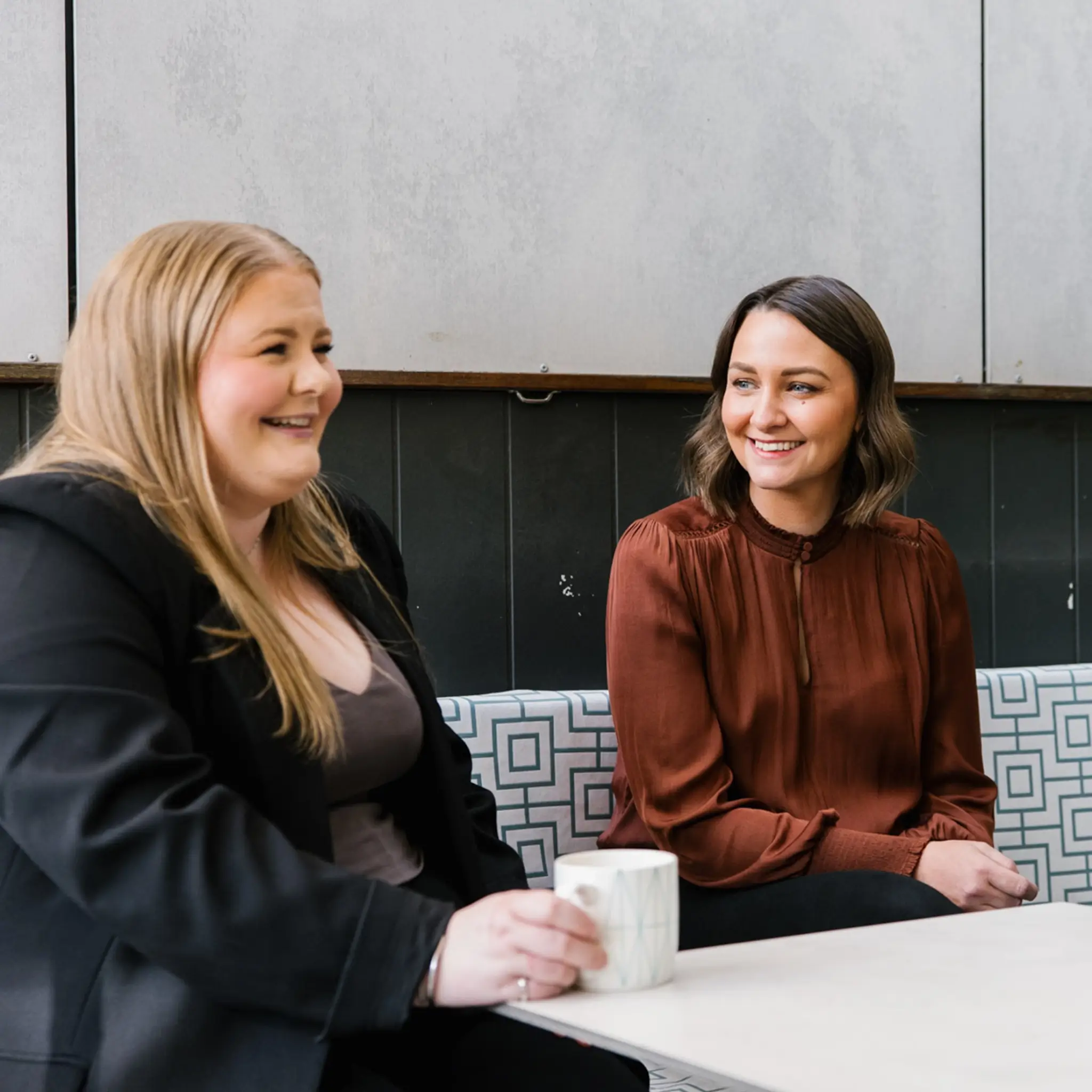 Two MyBudget colleagues are sitting together on a patterned bench in a cafe or office breakroom, smiling and talking. The woman on the left has long blonde hair and wears a black blazer over a dark top while holding a coffee mug. The woman on the right has shoulder-length brown hair and wears a rust-coloured silk blouse.
