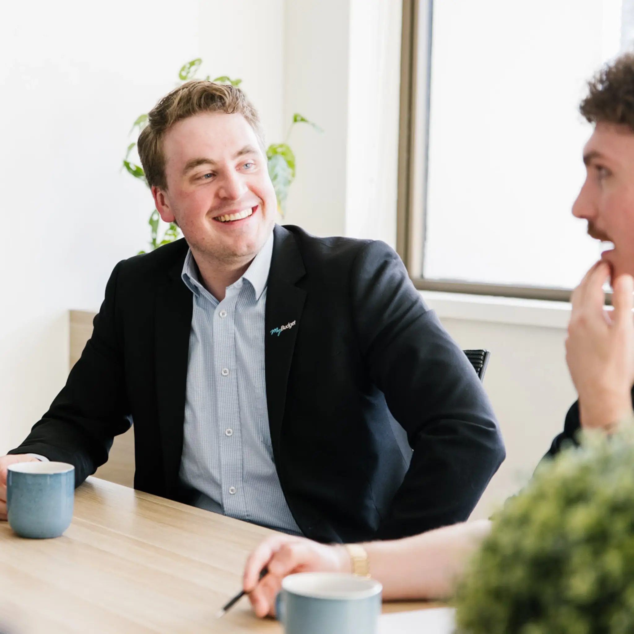 A smiling MyBudget employee in a black blazer and light blue button-down shirt sits at a light-coloured wooden table during a meeting. He is looking and laughing toward a colleague whose face is partially visible in the right foreground. A blue coffee mug sits on the table in front of him.