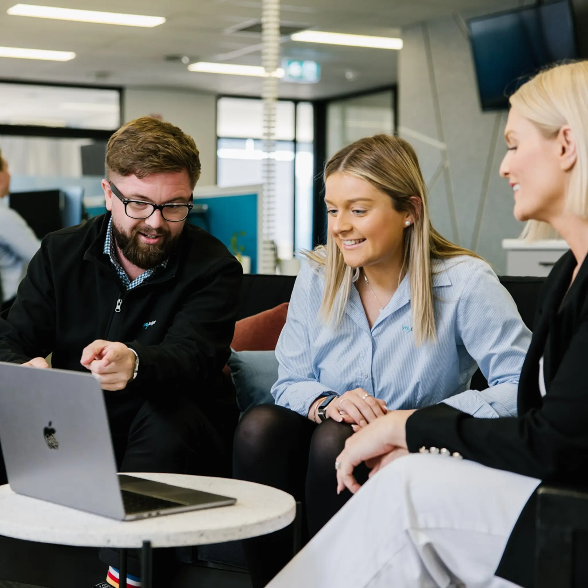 Three MyBudget colleagues—a man with glasses and a beard, a woman with blonde hair in a light blue shirt, and a woman in a black blazer and white pants—sit together in a modern office space, smiling as they look at a laptop screen on a small round table.