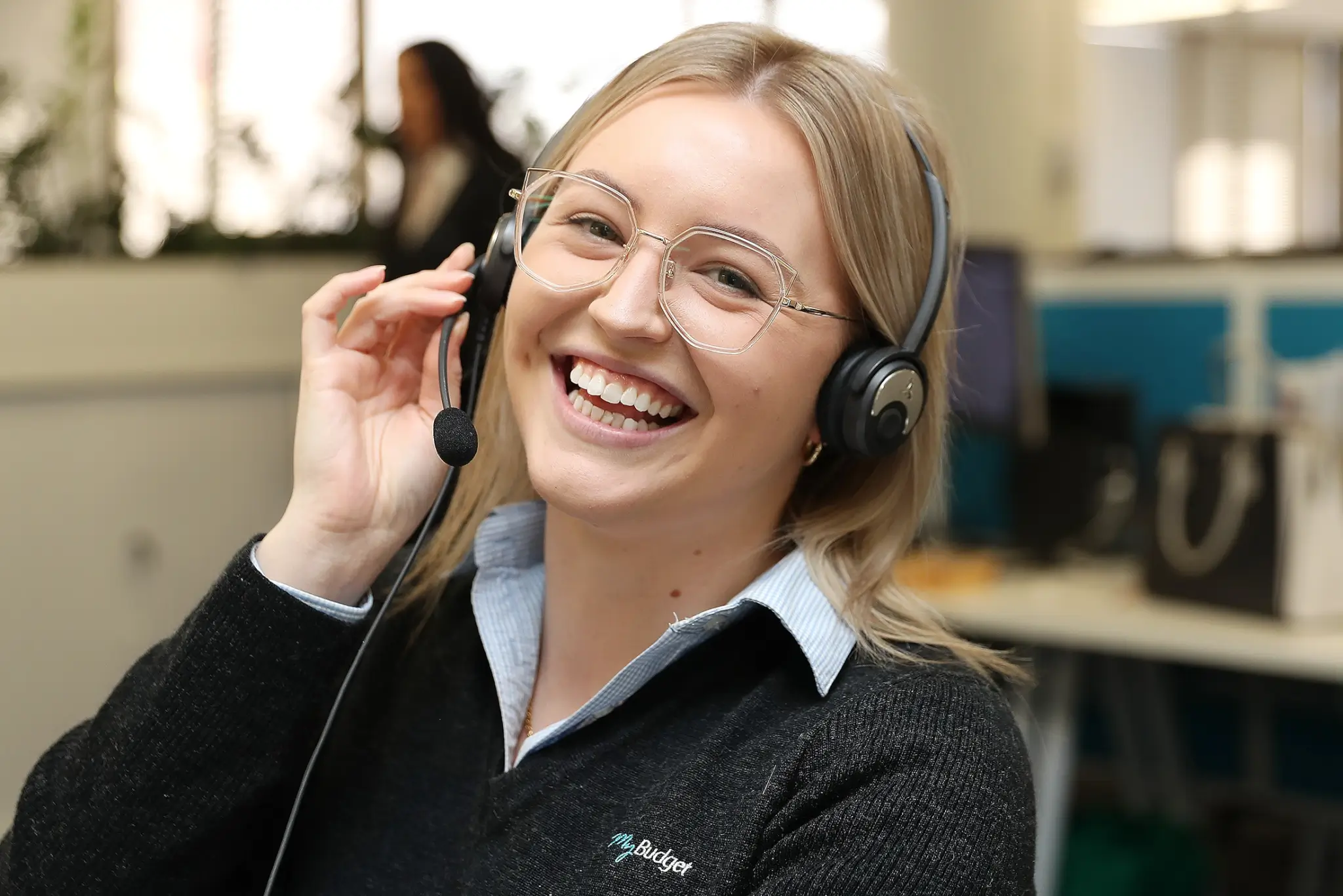 A young woman wearing glasses and a headset smiling brightly as she adjusts her microphone, wearing a black jumper with the MyBudget logo in an office environment.