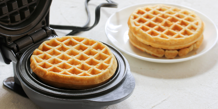 An opened mini waffle maker with a waffle in it and two more waffles stacked on a white plate next to it: Stocking fillers for women.