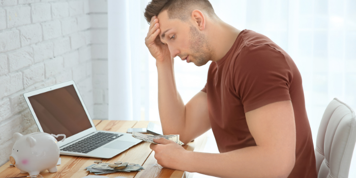 A young man stressed as he's sitting down hunched over money on a table with a piggy bank and an open laptop - can afterpay affect your credit rating?