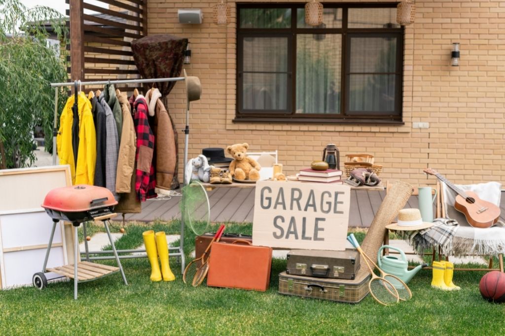 A garage sale set up on the outside of a cream brick house with a BBQ, guitar, old tennis racquets and suitcases, toys and a rack of clothes.