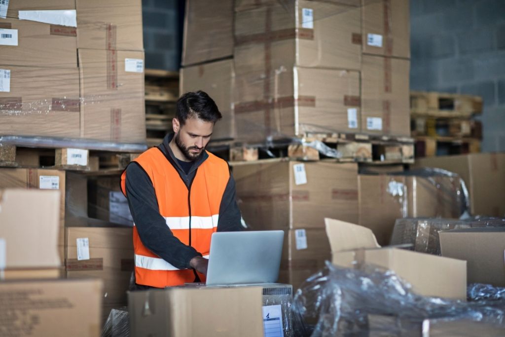 A man with black hair & beard wearing a high-viz vest typing on a laptop in a warehouse with pallets of boxes - Start-up costs for a business