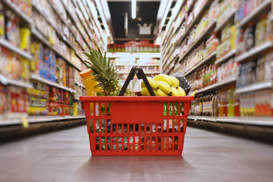 Red shopping basket with bananas, pineapple, juice, potatoes on the floor of an isle in a grocery store - tips on how to become better with money.