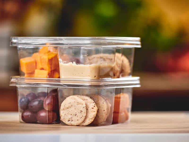 Eight transparent containers stacked on a kitchen bench with crackers, hummus, grapes and apricot squares in them