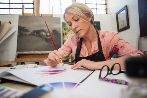 A woman wearing an apron in an art studio painting with other art on easels behind her in the background.