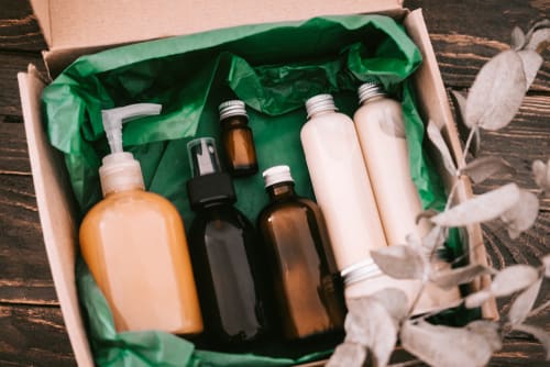 An open cardboard box with full soap dispensers, bottles and fragrances, with a flower stem to the side.