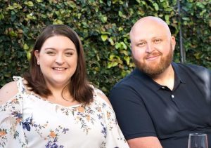 A woman wearing jeans and a jumper sitting at her dining table on the phone with her laptop in front of her while a young boy sits next to her. They’re smiling at each other.