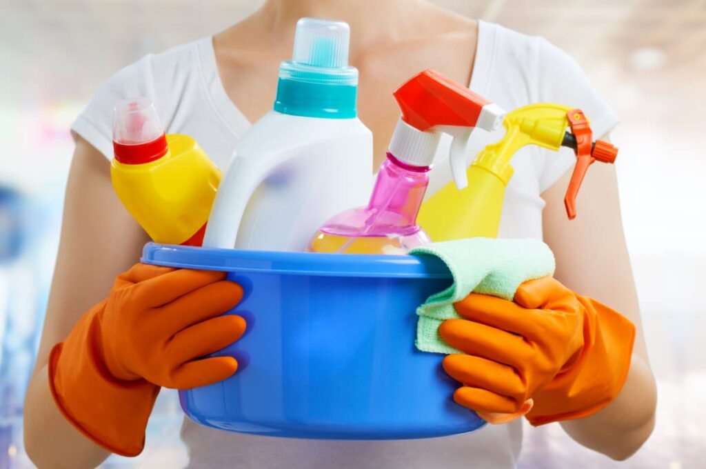 Person’s hands with orange gloves on holding up a blue bucket with cleaning sprays and a green cloth in it.