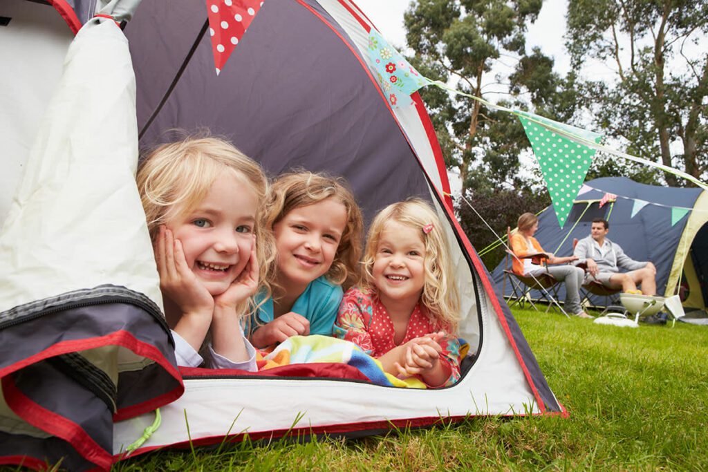 Three small children smile inside a camping tent, while their parents sit in fold-up chairs in the background.