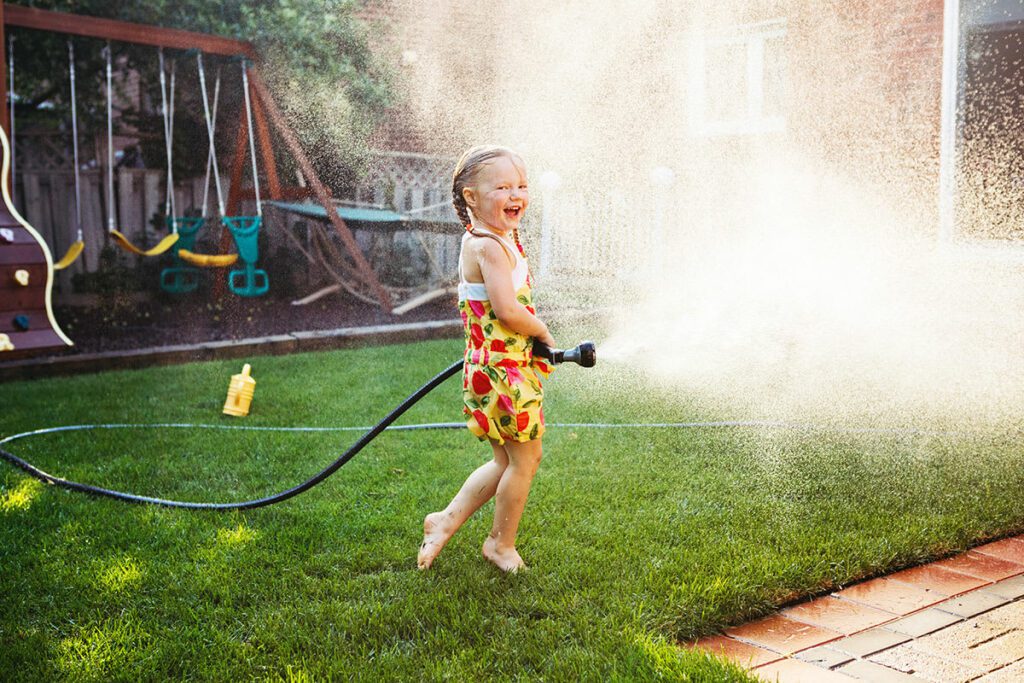 A small laughing girl blasts a hose around the backyard, sending water spraying everywhere.