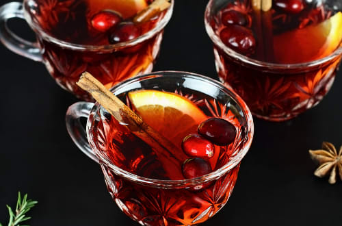 A glass of red Christmas punch filled with spices and fruits sits atop a black table.