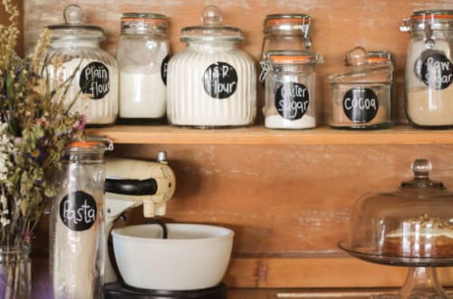 A wooden pantry is stocked full of flour, caster sugar, cocoa, raw sugar, pasta and mixing utensils.