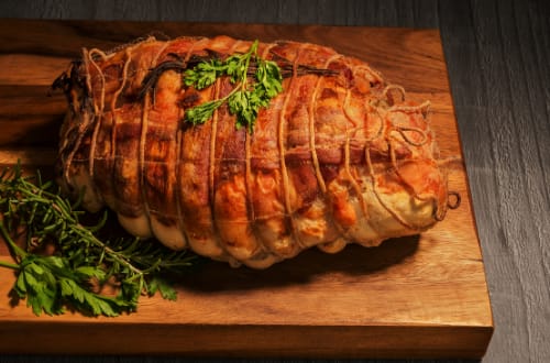 A roasted pork loin sits on top of a wooden chopping board and is accompanied by a range of herbs.