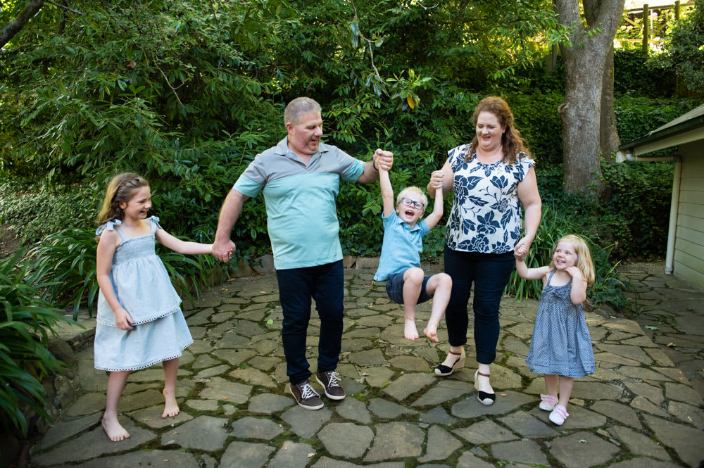 MyBudget clients Pete and Alyssa with three young children in a beautiful green garden. They are all holding hands and the parents are swinging their middle child between them.