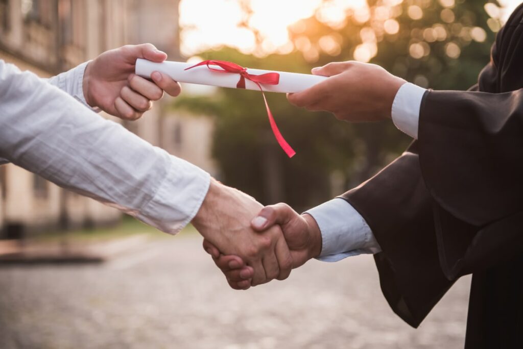 Close up photo of a person shaking hands with a graduate while passing over a diploma tied with a ribbon