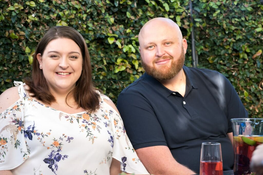 A married female and male couple in their late thirties sitting next to each other, smiling at the camera