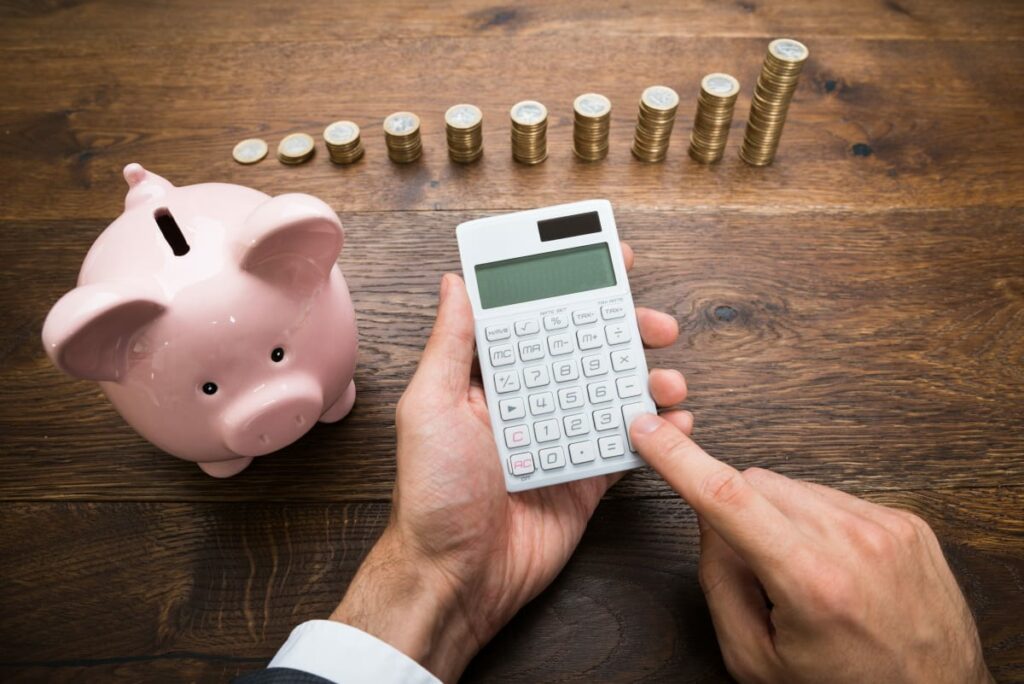 A person uses a calculator with coins stacked in incrementally bigger towers, with a piggybank sitting next to the coins.