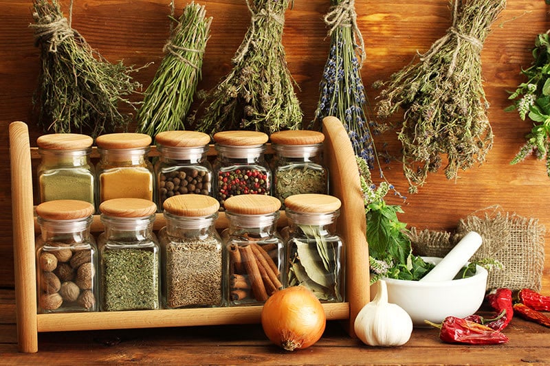 Wooden bench with a spice rack, onion, garlic, chilli and a mortar and pestle on it, herbs hanging behind and bench.