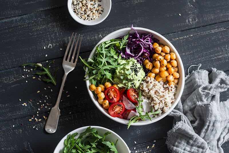 Black table with a fork, napkin and salad bowl on it with tomato, chickpeas, quinoa, red cabbage, rocket, avocado and red onion.