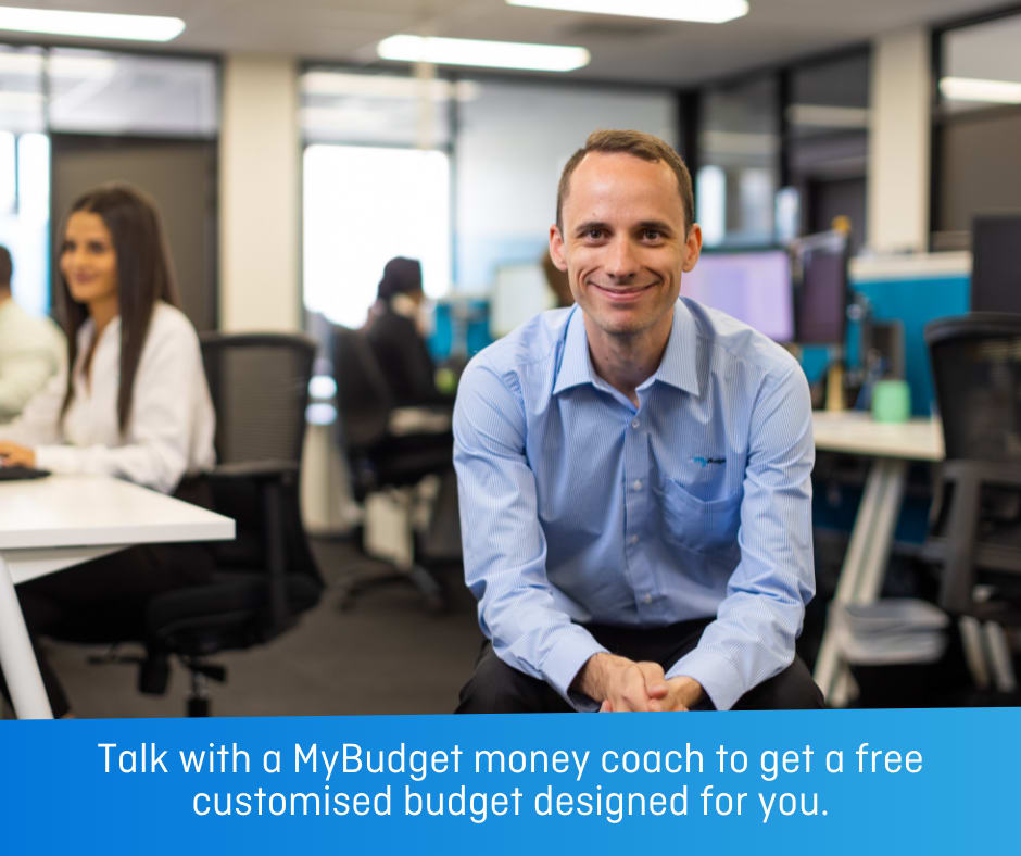 A man in a blue button up shirt smiles as he sits in an office. With text: Talk with a MyBudget money coach to get a free customised budget designed for you.