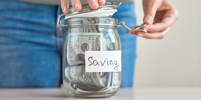 A person in jeans holding the lid of a clear jar with an American $100 bill and a white tag with "Saving" written on it.