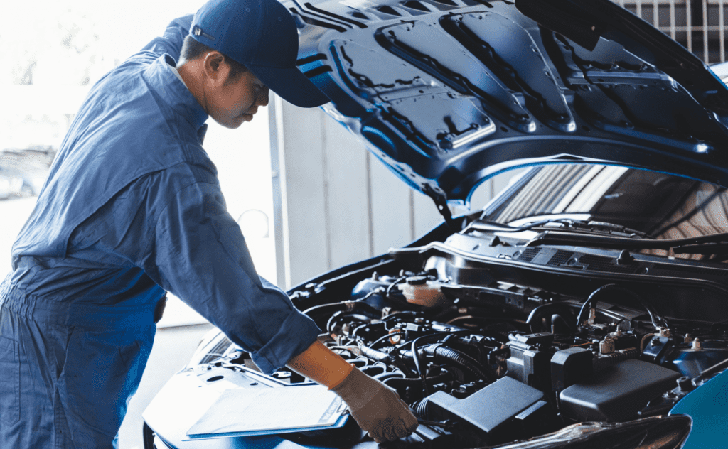 A mechanic in blue overalls lifts the hood of a blue car and inspects the engine.