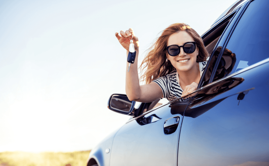 A woman holds car keys and smiles out the front window of her car. When to buy a new car vs used.